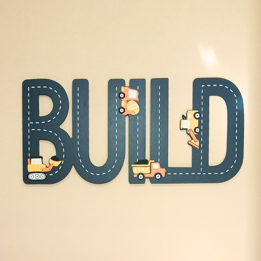 The word build cut out into a wooden sign, painted navy blue with little yellow construction trucks.